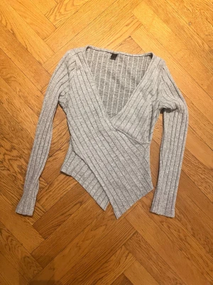 Grå ribbad omlottopp - Grå ribbad omlottopp med v-ringning och lång ärm. Toppen har en asymmetrisk nederkant och är figurnära i modellen. Perfekt att styla med jeans eller kjol för en trendig look.