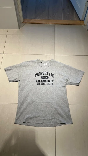Grå Gymshark t-shirt med tryck - Säljer en grå t-shirt från Gymshark med svart texttryck på bröstet där det står 'PROPERTY OF GSLC THE GYMSHARK LIFTING CLUB'. Klassisk passform med rund hals och korta ärmar. Perfekt för dig som gillar gym och streetwear.