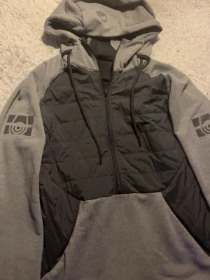 Grå och svart hoodie cardigan - Snygg hoodie cardigan i grått och svart med dragkedja framtill och quiltad front. Har tryck på ärmarna och huvan samt en gul detalj på baksidan av huvan. Justerbar huva med snören och mjukt material, perfekt för chill dagar. 