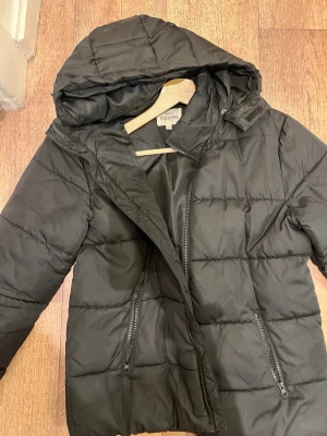 Svart pufferjacka från KappAhl - Säljer en svart pufferjacka från KappAhl med huva och dragkedja framtill. Jackan har två snedställda fickor med dragkedja och quiltad design. Perfekt för kalla dagar och riktigt skön att bära. Materialet är syntet och jackan har en klassisk, enkel look.