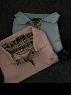 Burberry pikétröjor rosa & ljusblå - Två stilrena Burberry pikétröjor i rosa och ljusblått med klassiskt rutigt mönster på insidan av kragen. Tröjorna har broderad logga i guld på bröstet och är tillverkade i mjukt bomullsmaterial. Perfekta för en clean och trendig look.