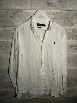 Vit linneskjorta från Polo Ralph Lauren - Klassisk vit skjorta i linne från Polo Ralph Lauren med lång ärm och knappar framtill. Skjortan har den ikoniska broderade loggan i marinblått på bröstet och en avslappnad passform. Perfekt till både jeans och chinos.