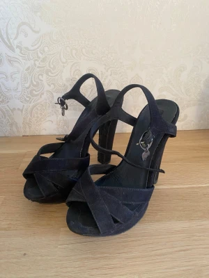 Svarta pumps med remmar 35 - Svarta pumps med höga klackar och korsade remmar framtill. Skorna har öppen tå och justerbar ankelrem med liten dekoration. Defekter finns