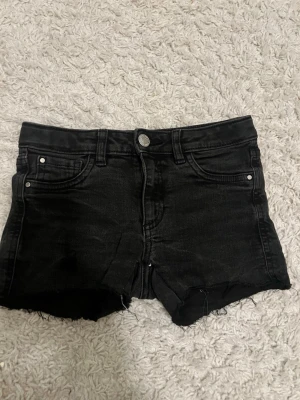 Svarta jeansshorts med rå kant - Säljer ett par svarta jeansshorts med rå, fransig kant och klassisk femficksdesign. Shortsen har knapp och dragkedja framtill samt bälteshällor. Perfekta för sommaren och ger en cool, avslappnad vibe.
