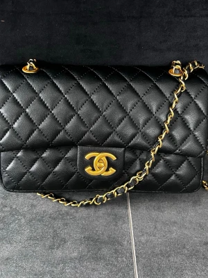 ChaneI svart axelväska - Säljer en klassisk svart axelväska från ChaneI i quiltat läder med guldfärgade kedjedetaljer. Väskan har en elegant rektangulär form och en ficka på baksidan. Perfekt för dig som gillar lyxiga accessoarer.