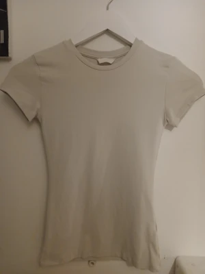 Beige t-shirt från H&M - En enkel och stilren beige t-shirt från H&M med rund halsringning och korta ärmar. T-shirten har en slim fit passform och är tillverkad i ett mjukt och stretchigt material som känns skönt mot huden. Perfekt basplagg till garderoben.💛Xs-S