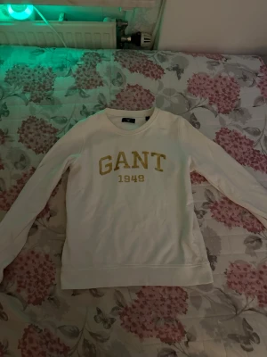 Vit sweatshirt från GANT strl XS - Säljer en vit sweatshirt från GANT med guldig text 'GANT 1949' på bröstet. Tröjan har rund halsringning och långa ärmar. Perfekt för en avslappnad och stilren look. Klassisk college-stil som aldrig går ur tiden.