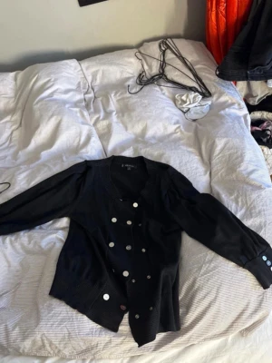 Anne klein kofta - Vintage Black Cardigan with Silver Buttons  This chic vintage black cardigan has a timeless and elegant design. It features double rows of beautiful silver buttons on the front, giving it a sophisticated and stylish look.  The cardigan has slightly shorter sleeves and a unique detail with small buttons at the ends of the sleeves, adding an extra touch of character. The structured design and classic color make it easy to style for both casual and more dressed-up outfits. 