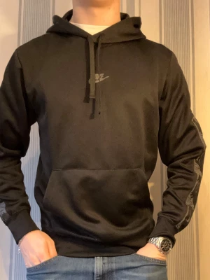 Svart Nike hoodie med logga - Svart hoodie från Nike med klassisk logga på bröstet och dragsko i huvan. Hoodien har en stor magficka och ribbade muddar vid ärmslut och midja. Perfekt för en avslappnad och sportig stil.