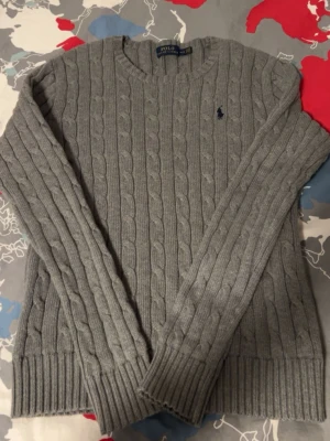 Grå kabelstickad tröja från Polo Ralph Lauren - Säljer en grå kabelstickad tröja från Polo Ralph Lauren med rund hals och diskret blå logga broderad på bröstet. Tröjan har långa ärmar och ribbade muddar vid ärmslut och nederkant. Perfekt för dig som gillar klassisk och stilren design. Den är helt ny aldrig använt.