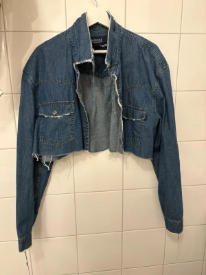 Cropped jeansjacka från Arizona - Säljer en blå cropped jeansjacka från Arizona med råa, fransiga kanter och två bröstfickor med knapp. Klassisk krage och långa ärmar. Perfekt för dig som vill ha en edgy och trendig look.