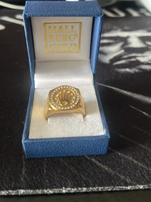 Guldfärgad ring med stenar - Snygg guldfärgad ring från Hallbergs Guld med en rund platta framtill, dekorerad med gnistrande stenar runt kanten och ett detaljerat emblem i mitten. Ringen har en bred och robust design som verkligen sticker ut.