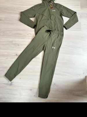 Grön träningsdress från Under Armour - Snygg grön träningsdress från Under Armour med dragkedja på jackan och elastisk midja på byxorna. Setet har vita loggor på både jacka och byxa samt svarta detaljer vid ärmslut och midja. Perfekt för gymmet eller löprundan.