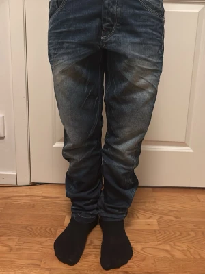 Blåa jeans med slitningar - Säljer ett par blå jeans med snygga slitningar och tvättade detaljer. Jeansen har klassisk femficksdesign, raka ben och kontrastsömmar på bakfickorna. Perfekta för en avslappnad och trendig look.
