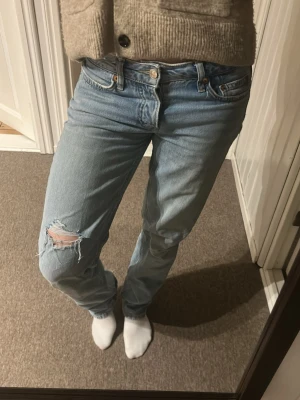 Ljusblå raka jeans med slitning - Säljer ett par ljusblå jeans med rak passform och låg midja. Jeansen har en snygg konstgjord slitning vid vänster knä Tillverkade i denim med knappgylf och bälteshällor. Perfekta för en avslappnad och trendig look.