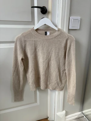 Beige stickad tröja från H&M - En stilren beige stickad tröja från H&M med rund halsringning och långa ärmar. Tröjan har ribbade muddar vid ärmslut och nederkant, vilket ger en snygg och enkel look. Perfekt för lager-på-lager och passar till det mesta.