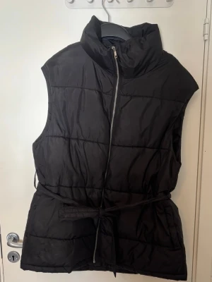 Svart dunväst från NLY Trend - Snygg svart dunväst från NLY Trend med hög krage, dragkedja framtill och ett matchande knytband i midjan. 