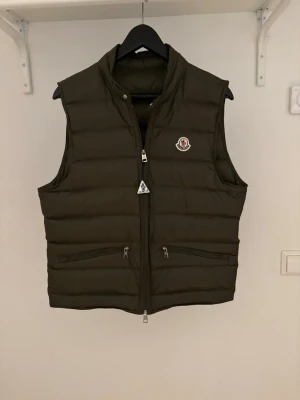 Mörkgrön dunväst från Moncler - Stilren mörkgrön dunväst från Moncler med hög krage och två dragkedjeförsedda fickor framtill. Västen har Moncler-logga på bröstet och quiltad design som ger en modern look. Perfekt för lager-på-lager och enkel att matcha till olika outfits.