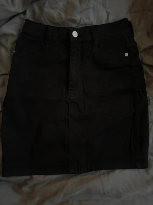 Svart jeanskjol  - Snygg svart jeanskjol från Bubbleroom med klassisk femficksdesign och knapp framtill. Som inte kommer till användning längre då den är för liten.  Kjolen är kort och har en rak passform, perfekt att matcha med en t-shirt eller hoodie. Gjord i ett stadigt bomullsmaterial med bälteshällor i midjan.