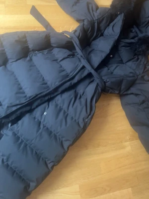 Svart lång dunjacka med huva - Snygg svart lång dunjacka med bälte i midjan och huva med pälskant. Jackan har quiltad design och stängs med både dragkedja och knappar. Perfekt för kalla dagar när du vill hålla dig varm och stilren.