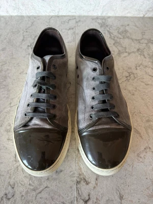 Lanvin Captoe  - Snygga sneakers i grå mocka med svart, glansig tå och svarta skosnören. Skorna har en vit sula och en stilren, låg profil som passar till många olika outfits. Perfekt för dig som gillar en clean och modern look. Pris 1699. Seriösa bud.