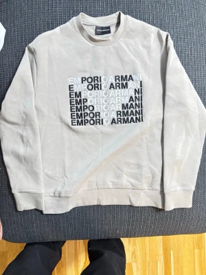 Beige sweatshirt från Emporio Armani - Säljer en beige sweatshirt från Emporio Armani med tryckt logga i svart och vitt på bröstet. Tröjan har rund halsringning, långa ärmar och ribbade muddar. Perfekt för en stilren och avslappnad look.
