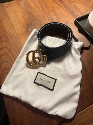 Svart skärp med GG-spänne från Gucci - Snyggt svart bälte från Gucci med ikoniskt dubbelt GG-spänne i guld. Bältet är gjort i slätt skinn och har en klassisk, stilren design som passar till många outfits. Perfekt accessoar för att lyfta din look.  Säljer då det är för litet! Storlek: 90 cm