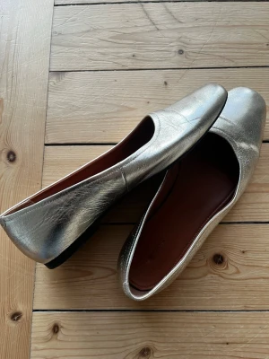 Guldiga ballerinaskor från Vagabond - Snygga ballerinaskor i metallic guld från Vagabond. Skorna har en klassisk, rundad tå och är tillverkade i skinn med en mjuk innersula. Perfekta för dig som vill sticka ut med en trendig och stilren look.