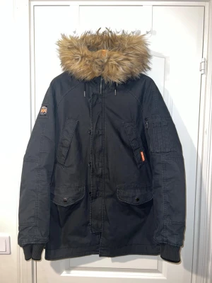 Superdry parkasjacka - Svart parkasjacka från Superdry med stor huva och fuskpäls. Jackan har flera fickor framtill, ärmslut med muddar och patch med brittisk flagga på ärmen. Perfekt för kalla dagar och riktigt snygg streetstil.