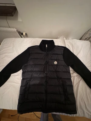 Svart dunväst från Moncler - Snygg svart cardigan från Moncler med stickade svarta ärmar och hög krage. Västen har dragkedja framtill, två fickor med dragkedja och Moncler-logga på bröstet. Perfekt för lager-på-lager och streetstyle.