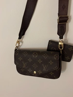 Superdin brun axelväska - Snygg axelväska  i klassiskt brunt monogrammönster med guldfärgade detaljer. Väskan har en bred axelrem och en mindre avtagbar plånbok framtill. Väskan är oanvänd och i supergott skick. 