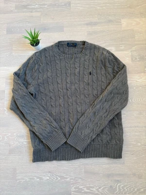 Grå kabelstickad tröja Polo Ralph Lauren - Kabelstickad grå tröja från Polo Ralph Lauren med rund halsringning och klassisk marinblå logga broderad på bröstet. Tröjan har ribbade muddar vid ärmslut och nederkant. Perfekt för dig som gillar tidlös och stilren design.