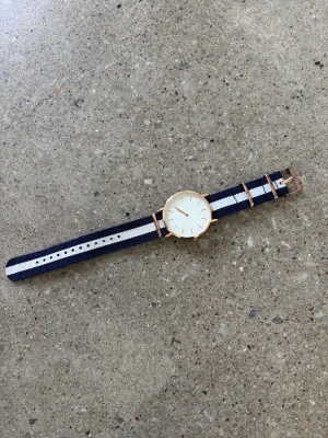 Klocka  - Stilren klocka med vitt ur och randigt band i marinblått och vitt. 