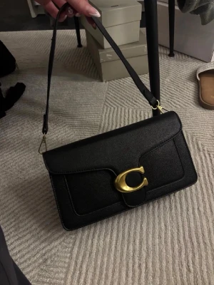 Svart axelväska - Svart axelväska i strukturerat material med guldfärgad spänndetalj framtill. Slank och stilren modell med lockstängning och justerbar axelrem. Insida med flera fack som gör det enkelt att hålla ordning. Funkar lika bra till vardag som kväll.