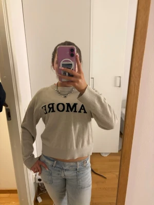 Beige sweatshirt med svart text - Trendig beige sweatshirt med svart texten 'AMORE' över bröstet. Croppad modell med rund halsringning och långa ärmar. Perfekt till jeans för en avslappnad look.