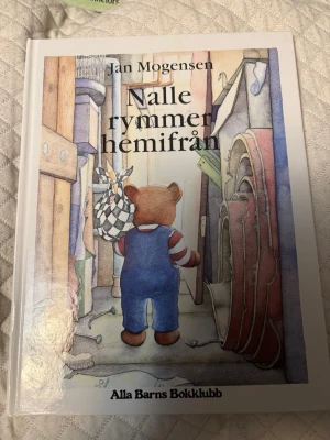 Nalle rymmer hemifrån - Jan Mogensen - Säljer boken 'Nalle rymmer hemifrån' av Jan Mogensen. Det är en illustrerad barnbok med hårda vita pärmar och färgglada teckningar på framsidan. Perfekt för dig som gillar mysiga berättelser om äventyr och vänskap.