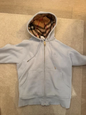 Ljusgrå hoodie från Burberry, den är ifrån burbery outlet från Paris, har använt den några gånger och är i bra skick men det är lite smuts på högra ärmen där nere vid handen men annars ser den bra ut - Snygg ljusgrå hoodie från Burberry med dragkedja och klassiskt rutigt foder i huvan. Hoodien har två stora fickor framtill och guldfärgad dragkedja med Burberry-logga. Perfekt för dig som gillar exklusiv streetwear och vill sticka ut med ikoniska detaljer.