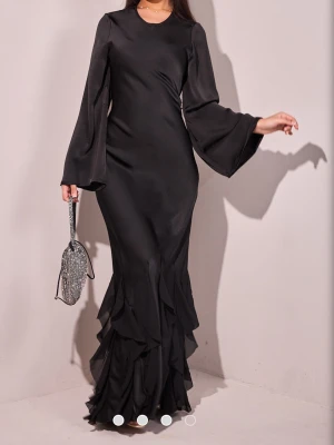 Svart långklänning med volangärm - Elegant svart långklänning i ett glansigt material med vida, långa ärmar och volangdetaljer längst ner. Klänningen har en rund halsringning och figurnära passform upptill. Perfekt för dig som vill sticka ut med en stilren look. Storleken är s/m men går mer mot m. Jag har aldrig använt denna då jag inte ofta använder svarta kläder 💞