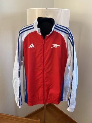 Adidas Arsenal vind/träningsjacka  - Säljer en snygg Arsenal vind/träningsjacka från Adidas Storlek M. Det är alltså 2 jackor i 1. Om man vänder den ut och in så blir det en annan i annan färg. Så på det ena hållet så är jackan röd med vita ärmar med blå adidas ränder och blå detaljer längs sidorna. O åt andra hållet så är jackan svart med lite vita, röda och gröna detaljer. Jackan har en full dragkedja framtill, ståkrage och elastiska muddar. Tillverkad i lätt och funktionellt material, perfekt för all typ av användning. 