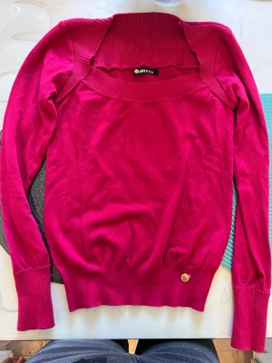 Mörkrosa stickad tröja Artistry - Snygg mörkrosa stickad tröja från Artistry med bred ribbad krage och långa ärmar. Tröjan har en figurnära passform och ribbade muddar vid ärmslut och nederkant. Perfekt för dig som vill ha en färgstark och bekväm stickad topp.