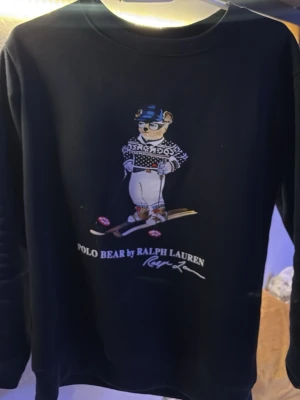 Svart Polo Bear hoodie Ralph Lauren - Svart hoodie från Polo Ralph Lauren med ikoniska Polo Bear-trycket framtill där björnen åker skidor. Klassisk rund halsringning och mjukt material på insidan. Perfekt för dig som gillar streetwear och vill ha en unik detalj i garderoben.