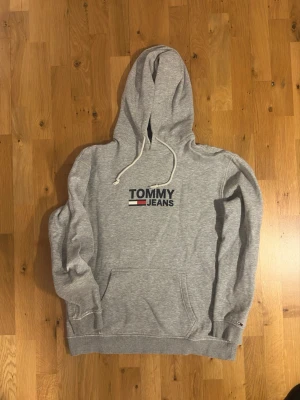 Grå hoodie från Tommy Jeans - Snygg grå hoodie från Tommy Jeans med klassisk logga på bröstet i blått, rött och vitt. Hoodien har huva med dragsko och en stor magficka framtill. Perfekt för en avslappnad och trendig stil.