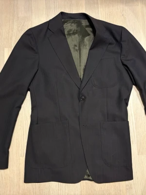 Tiger Of Sweden kavaj med kritsträck - Oanvänd Stilren mörkbrun kavaj från Tiger Of Sweden med diskreta kritsträck, tre utanpåliggande fickor och enkelknäppning. Kavajen har fyra knappar vid ärmsluten och är helfodrad med mörkgrönt innerfoder. Passar perfekt till en dressad look och har en klassisk, rak passform.