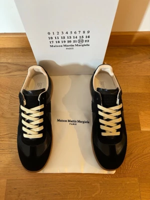 Maison Margiela svarta sneakers - Svarta sneakers från Maison Martin Margiela med vita kontrasterande skosnören och beige innersula. Klassisk låg modell med stilren design och gummisula. Materialmix av mocka och läder för en lyxig känsla.