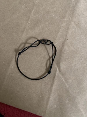 Svart armband från Valentino - Snyggt svart armband från Valentino med justerbart band i textil och en metallbricka med det ikoniska V-logotypen. Perfekt accessoar för dig som gillar stilrena detaljer och vill addera lite lyx till din look.