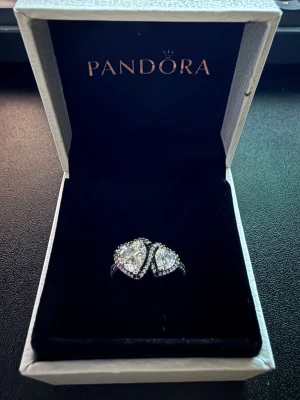 Pandora silverring med hjärtformade stenar - Säljer en snygg silverring från Pandora med två stora hjärtformade klara stenar och mindre stenar runtom. Ringen har en elegant och glittrig design som verkligen drar blickarna till sig. Perfekt för dig som gillar smycken med det lilla extra.