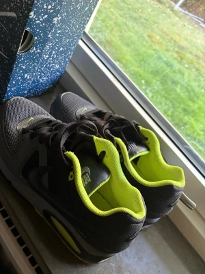 Nike Air Max grå/svarta sneakers - Säljer ett par Nike Air Max sneakers i grått och svart med neongula detaljer och innersula. Skorna har en klassisk låg siluett, mesh och syntetmaterial för bra andningsförmåga och komfort. Swoosh-logga på sidan och synlig Air Max-dämpning i sulan.