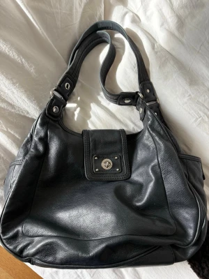Marc by Marc Jacobs väska - Säljer nu min fina Marc by Marc Jacobs-väska i svart läder och silverdetaljer i mycket fint och välbevarat skick. En klassisk och rymlig modell som passar både till vardag och arbete. Jag har anvant den som skolväska, och den rymmer utan problem A4-block och en laptop. Säljs med tillhörande dammpåse. Ett av magnetfästena på en av sidfickorna har något svagare magnetfunktion, men i övrigt är allt som det ska.