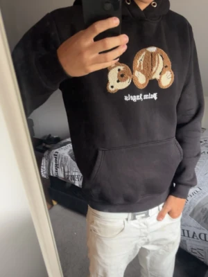 Svart Palm angels  hoodie med nalletryck - Säljer en svart hoodie med huva och stor magficka. På bröstet finns ett broderat motiv av en nalle och texten 'Palm Angels' i vitt. Hoodien har ribbade muddar och en avslappnad passform. Perfekt för dig som gillar streetwear och vill sticka ut.