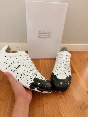 Maison Margiela GATs - Maison Margiela sneakers i vitt läder med svart färgstänk och unik målad tå. Klassisk låg modell med vita skosnören och detaljerad sula. Perfekt för dig som gillar statement-plagg och vill sticka ut med en kreativ och modern look.